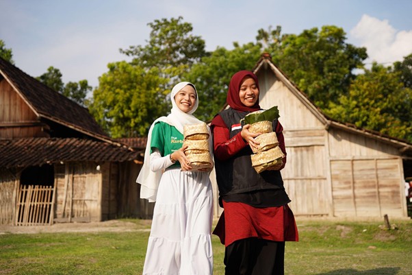 kurbanroisojk9 dompet dhuafa Two smiling women in hijabs carry stacked loaves of bread outdoors in a village setting with wooden huts and trees nearby.