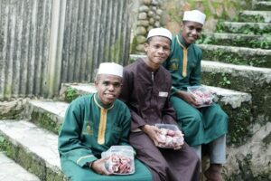 Three young men in traditional attire sit on stone steps, each holding a plastic tub of meat with smiles on their faces.