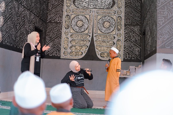 kurbantokopedia6 dompet dhuafa A young boy in an orange robe with a white cap speaks into a microphone on a stage, while a kneeling child and a standing woman clap nearby, with Arabic calligraphy on the backdrop and audience in the foreground.