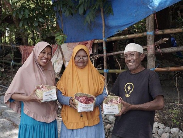 Three smiling people stand outdoors, each holding small baskets of fruit or produce in front of a wooden fence. The two women wear hijabs and colorful clothing, and a man in a white cap stands with them.