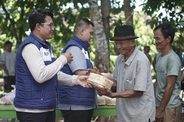 kurbanupzbazma11 dompet dhuafa Volunteers in blue vests hand a wrapped gift to an elderly man wearing a hat in a sunny park.
