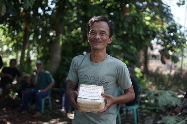 kurbanupzbazma12 dompet dhuafa Man in a teal t-shirt holds a woven gift basket with a label, outdoors in a park with trees and spectators in the background.