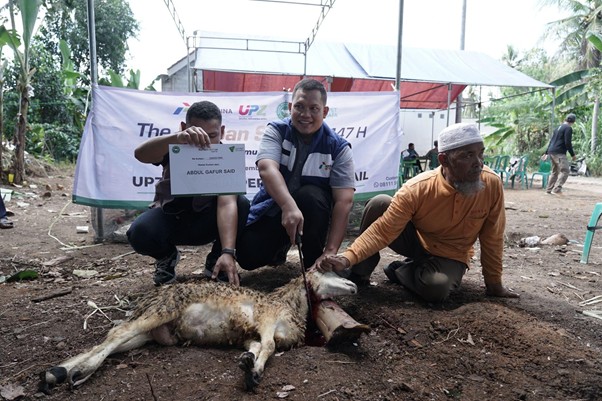 kurbanupzbazma3 dompet dhuafa Three men kneel around a dead animal on the ground at an outdoor community event, one holding a sign with a banner in the background.