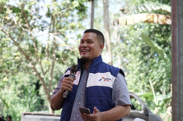 kurban upz bazma 7 dompet dhuafa Smiling man in a blue and white vest speaks into a microphone outdoors, holding a phone, with trees in the background.