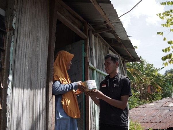 kurban upz bazma 9 dompet dhuafa Young man hands a wrapped box to a woman in an orange headscarf at the doorway of a weathered corrugated‑metal house.