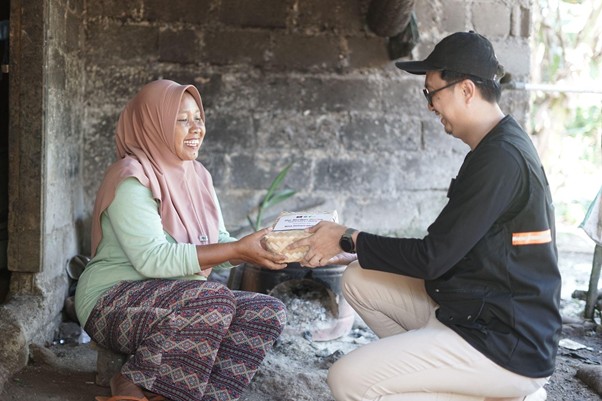 Man wearing a cap hands a box of food to a smiling woman in a hijab, seated in a rustic doorway.