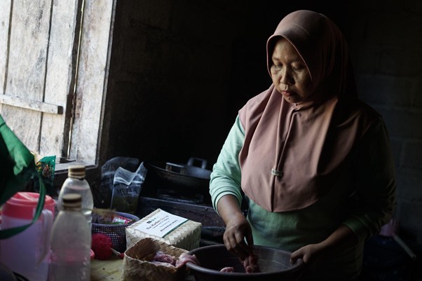 kurban di lampung timur 5 dompet dhuafa Woman wearing a hijab chops meat in a bowl on a worn kitchen counter near a sunlit window in a dim room.