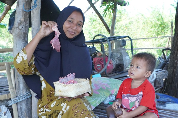 Woman in a navy hijab shows pink meat chunks from a woven basket to a young boy outdoors near trees and farming gear.