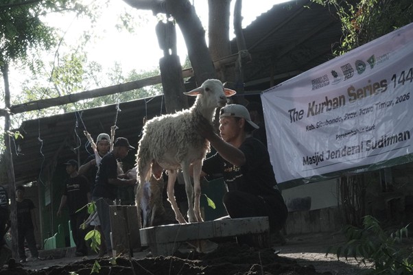 Man holding a white sheep on a raised platform during a Kurban Series event at Masjid Jenderal Sudirman; banner and onlookers visible in the background.
