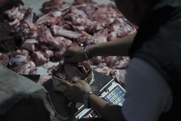 Butcher weighing a cut of meat on a scale with piled meat in the background.