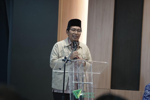 Male speaker wearing a beige jacket and black hat giving a talk at a clear podium with a microphone in a conference room, with a logo on the podium.