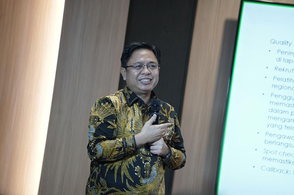Smiling man in a batik shirt giving a presentation, standing beside a projected slide with bullet points.