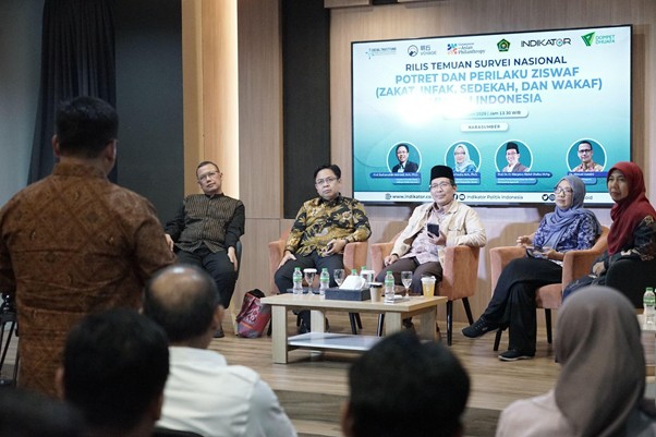 Six panelists seated on a stage discussing at a seminar, with an audience in the foreground and a blue banner behind them.|The scene shows a panel discussion with six speakers on stage and attendees watching from the audience, against a blue banner backdrop.