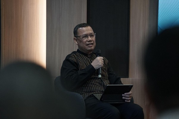 Man seated on a stage or panel, speaking into a handheld microphone while holding a tablet, audience blurred in the foreground.