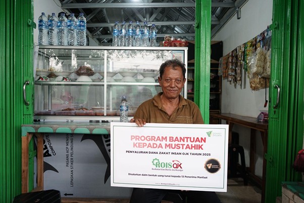 Smiling man in a brown shirt sits behind a small shop counter, holding a sign about aid to mustahik from ROISOK.