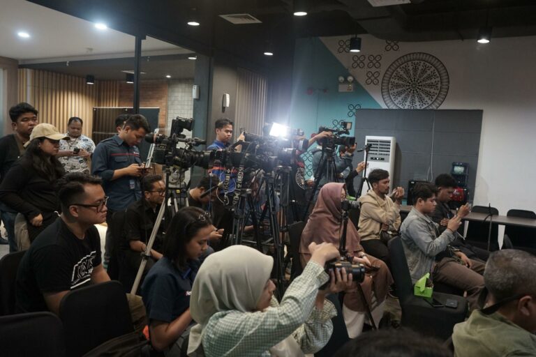 Group of journalists and photographers with multiple cameras filming a press briefing in a conference room.