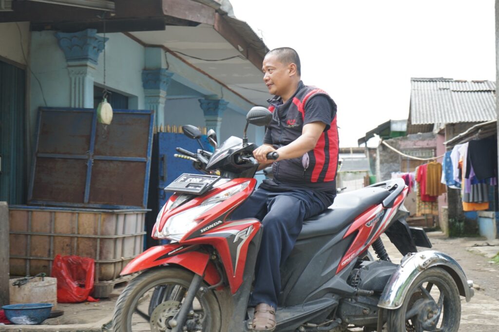 photo20220224120740 dompet dhuafa Man wearing a red and black vest sits astride a red Honda motorcycle on a narrow street with houses and laundry hanging to dry in the background, looking to the right of the frame.