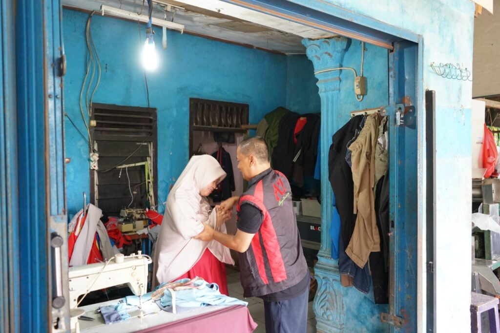 photo202202241207411 dompet dhuafa A tailor helps a woman in a light hijab adjust clothing inside a bright blue shop with a sewing machine and hanging clothes nearby.