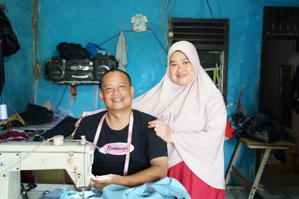 photo20220224120744 dompet dhuafa A man sits at a sewing machine with a measuring tape around his neck, smiling, as a woman in a light hijab stands behind him with hands on his shoulders in a blue room workshop