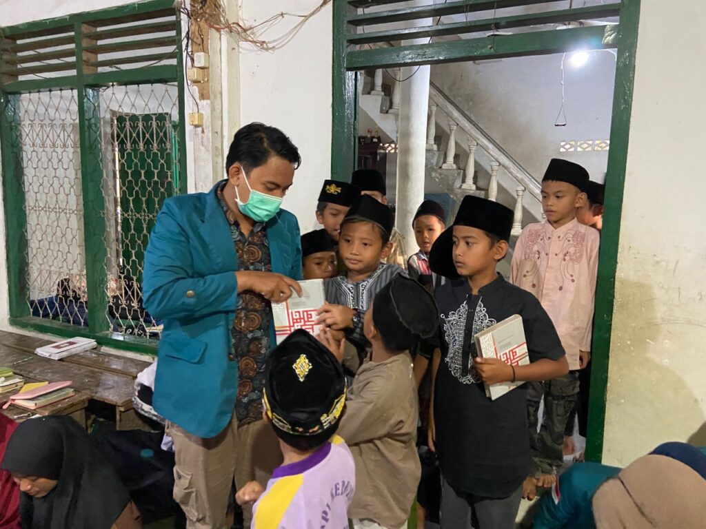 photo20220224175029 dompet dhuafa Man in a teal blazer and mask hands out items to a line of smiling children in traditional attire inside a doorway.