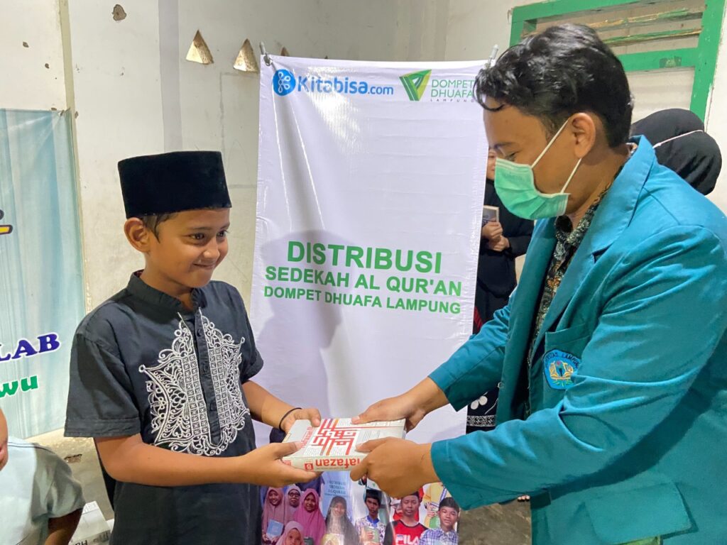 photo20220224175030 dompet dhuafa Young boy in a black cap receives a Qur'an box from a man in a teal coat and green mask at a charity distribution event, with a banner that reads 'Distribusi Sedekah Al Qur'an' in the background.