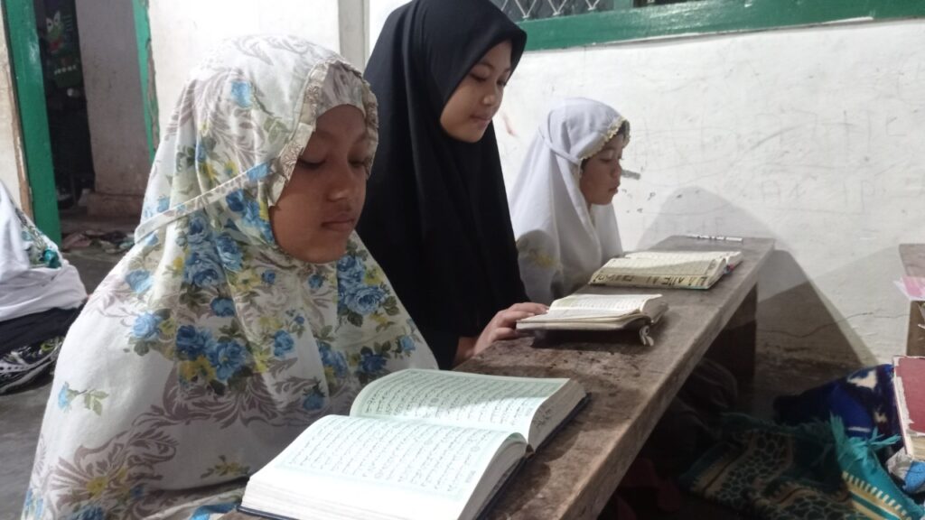 photo20220224175031 dompet dhuafa Three girls wearing hijabs sit at a wooden bench reading open books outdoors, focusing on their texts.
