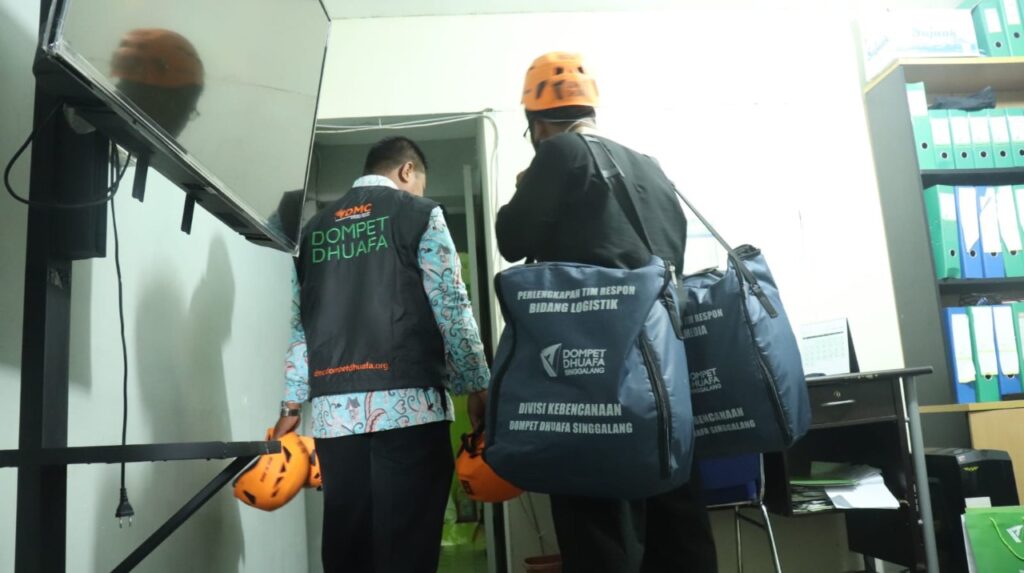 Two disaster-relief volunteers in orange helmets and Dompet Dhuafa vests carry large relief bags into a doorway.