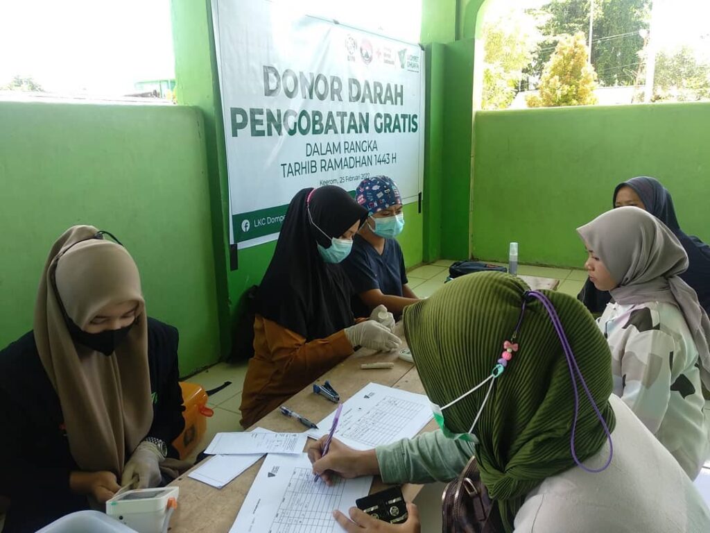 photo20220304085122 dompet dhuafa People at a donation table signing forms for a free blood treatment event, with a green banner reading 'Donor Darah Pengobatan Gratis' in the background, all wearing masks and hijabs.