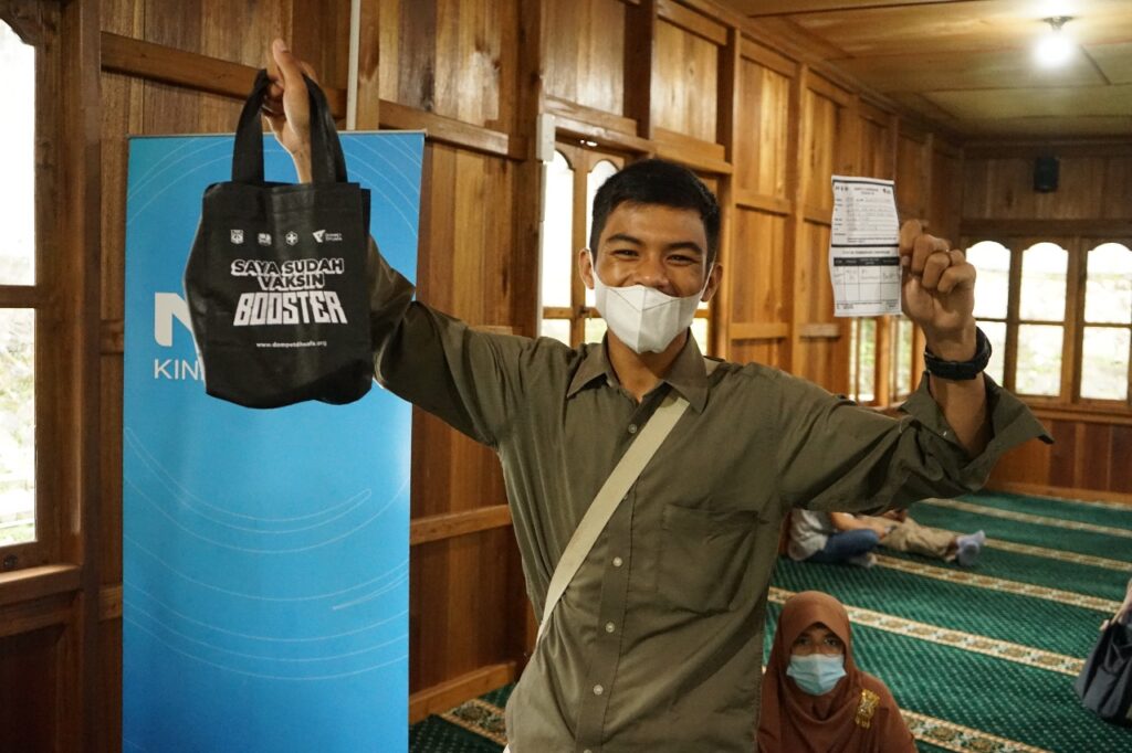 Man in a mask raises a black tote bag labeled 'Saya Sudah Vaksin Booster' in one hand and holds a vaccination card in the other, indoors with a blue banner nearby.