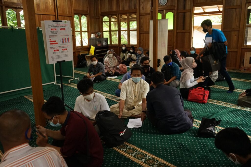 Group of people wearing masks sit on a green patterned carpet in a wooden hall, a health information poster on a stand nearby.