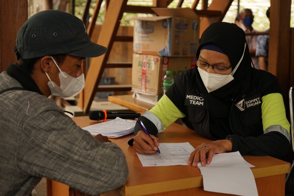 Medical volunteer in hijab and mask signs paperwork at a table while another masked person assists; boxes and supplies in the background at a relief station.