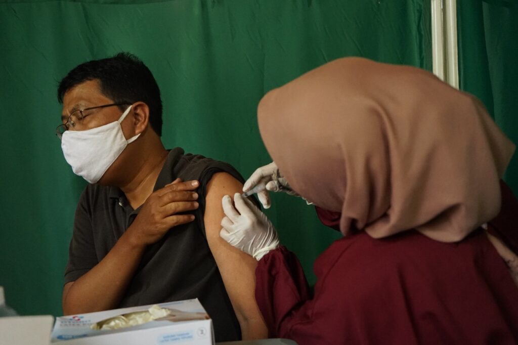 Man wearing a white mask receives a vaccination in the left upper arm from a nurse wearing a beige headscarf, in a clinic with a green backdrop.