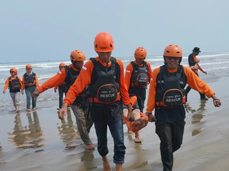 Group of rescue workers in orange uniforms and helmets walk along a sandy beach, some carrying a child between them in shallow surf.