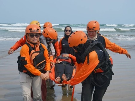 Group of lifeguards or rescuers in orange life jackets and helmets carry a person on a stretcher toward the beach, with waves in the background.