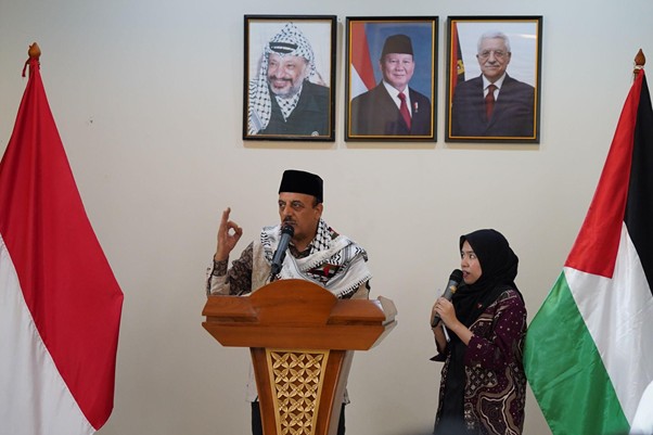 peringatanharinakbake784 dompet dhuafa A man wearing a kufi and keffiyeh speaks at a wooden podium with a microphone, while a woman in a hijab holds another microphone beside him; red-and-white flag on the left, Palestinian flag on the right, and three framed portraits of leaders on the wall behind them.