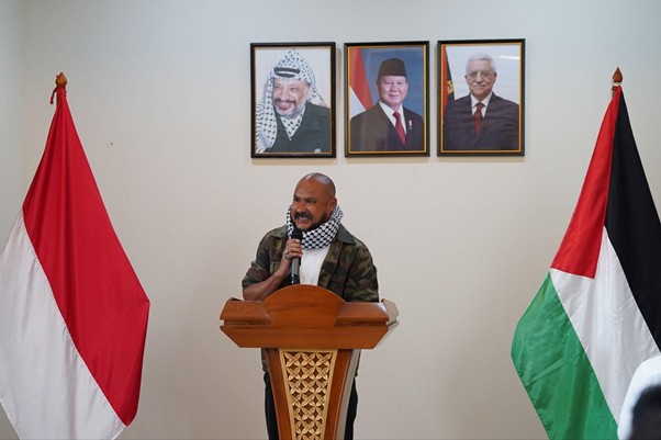 peringatanharinakbake785 dompet dhuafa Bald man in camouflage jacket speaks at a wooden podium, flanked by Indonesian and Palestinian flags, with three framed portraits of world leaders on the wall.