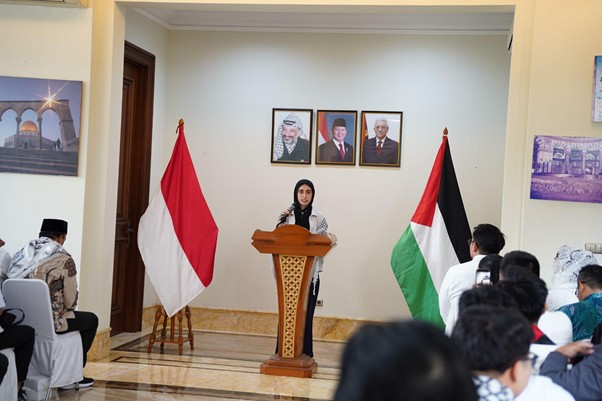 peringatanharinakbake786 dompet dhuafa Woman at a wooden podium delivering a speech in a hall, flanked by Indonesian and Palestinian flags, with portraits of leaders on the wall and a seated audience.