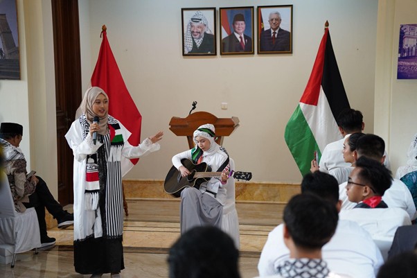 peringatanharinakbake789 dompet dhuafa Two women perform on a stage: one sings into a microphone while the other plays guitar, with Palestinian flags and portraits in the background and an audience watching.