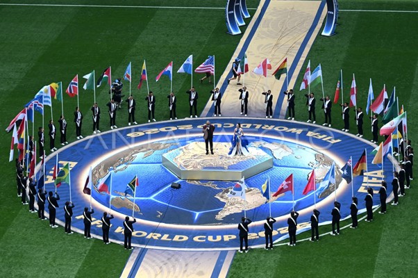 Large circular stage on a soccer field with a world-map design, surrounded by national flags on poles and uniformed attendants, as two people stand on a central podium during a ceremony.