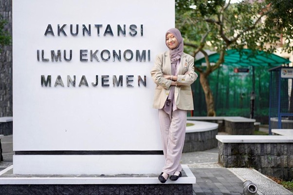 Woman in a hijab and beige blazer stands next to a white pedestal sign that reads 'AKUNTANSI ILMU EKONOMI MANAJEMEN' on a campus plaza.