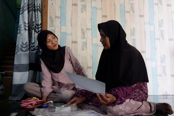 radha heryani putri 2 dompet dhuafa Two women wearing hijabs sit on the floor, looking over papers together in a casual indoor setting.