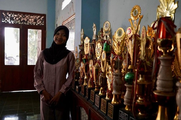 radha heryani putri 4 dompet dhuafa Young woman wearing a hijab stands beside a row of gold trophies on a shelf in a dimly lit room.