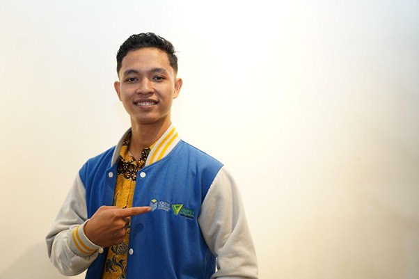 Smiling man in a blue varsity jacket points to a logo on his chest.",