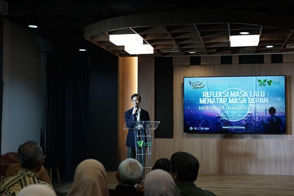 Speaker at a podium delivering a presentation on a stage; audience seated and facing forward with a blue title slide on a screen behind him, reading 'Refleksi Masa Lalu Menata Masa Depan'
