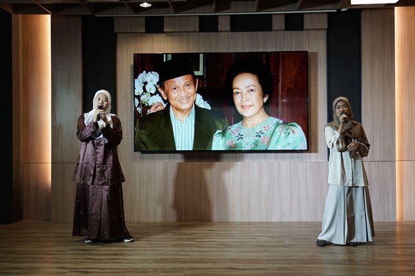 refleksi jati diri 2 dompet dhuafa Two women in hijabs speak on a wooden stage, with a large screen showing an elderly couple in the background.
