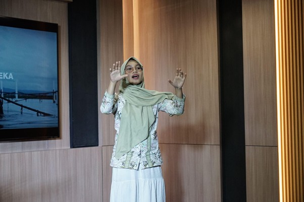 Woman in a beige hijab and glasses speaks with her hands raised during a talk in a wood-paneled conference room, with a screen visible on the left.