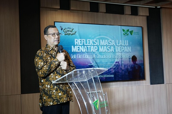 refleksi jati diri 4 dompet dhuafa Man wearing a batik shirt speaks into a microphone at a transparent podium on a stage, with a projector screen behind displaying Indonesian text.