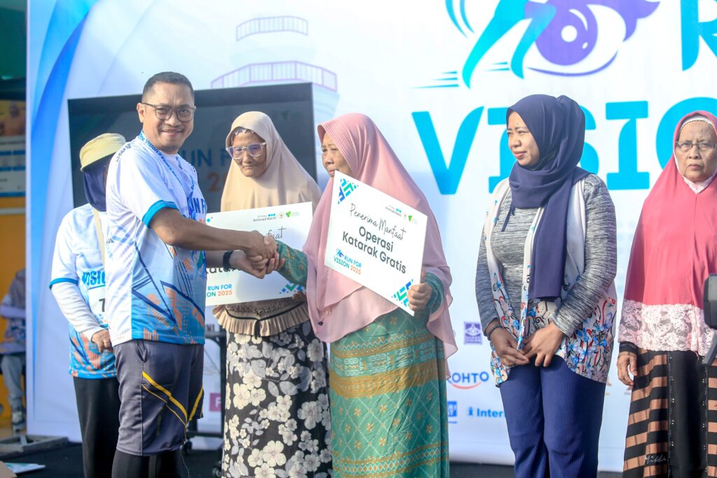 Man shakes hands with a woman in a hijab as others smile, holding signs advertising a free cataract operation at a health charity event on a stage backdrop