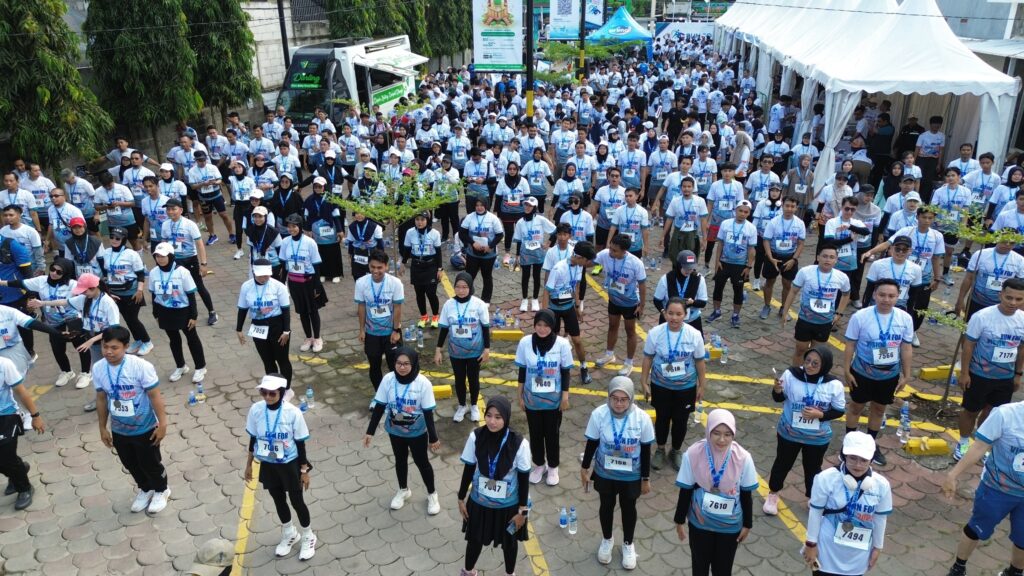 Large crowd of runners in light blue shirts at a charity run, bib numbers visible, with tents and a van along a paved street in the background.