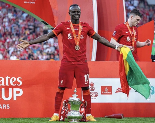Liverpool player in red kit with medal, arms outstretched on stage beside large trophy at Emirates Cup podium (sports award moment).